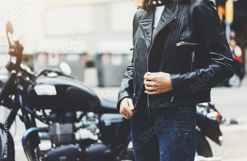 Girl unfastens black leather jacket on background motorcycle in sun flare city, hipster biker female hands closeup, motorbike street lifestyle, traveler planing bike route in summer holiday concept