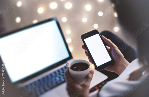 Hipster girl using computer and cup coffee in home atmosphere, person holding blank smartphone on background glow illumination, female hands texting on relax glitter bokeh, mockup templates, blur