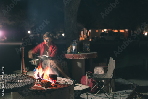 Woman cooking with fire wood and braai equipment by night. Tent and chairs in the foreground. Adventures in african national parks. Toned image.