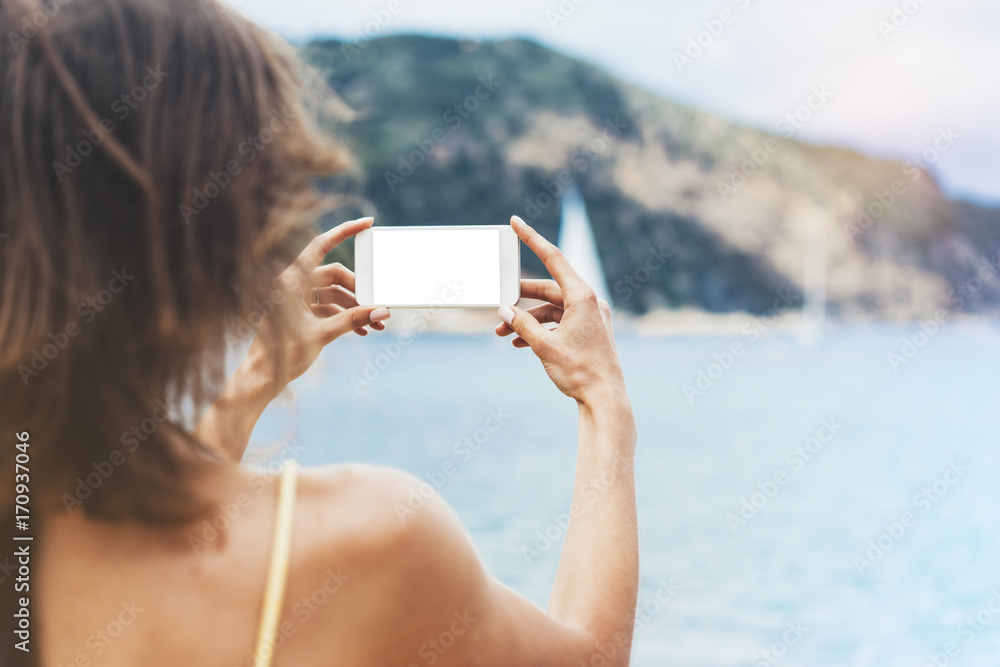 Hipster girl photograph on smart phone gadget in sand coastline, mock up of blank screen. Traveler using in female hand mobile on background seascape horizon. Tourist and blue sun ocean, lifestyle