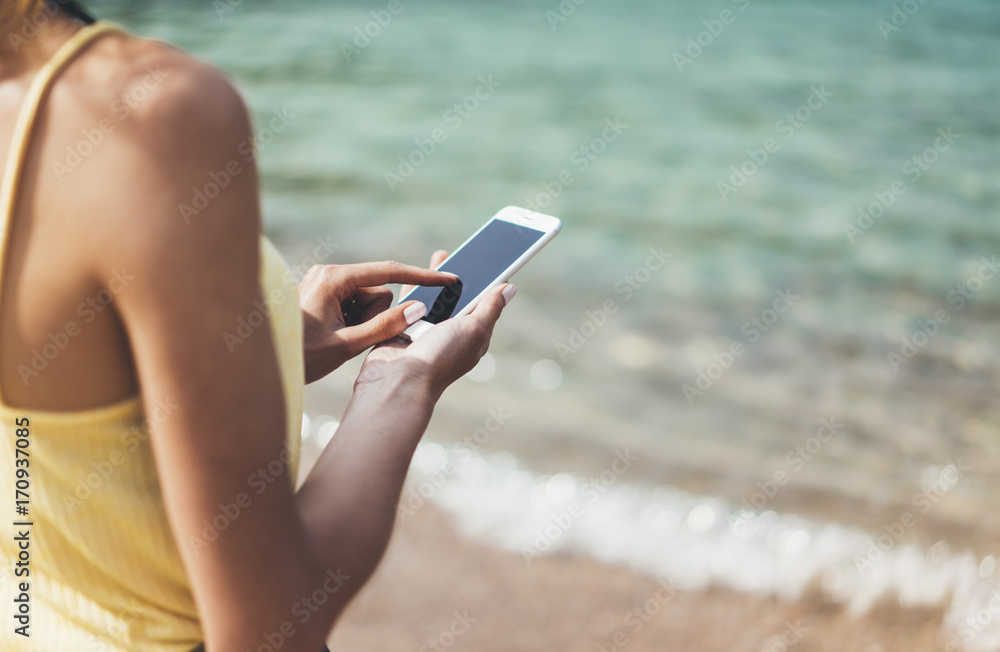 Hipster girl hold on smart phone gadget in sand coastline. Traveler using in female hand mobile on background beach seascape horizon. Tourist look on blue sun ocean, summer lifestyle