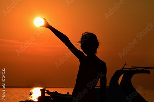 Silhouette of small girl holding sun during sunset above marina, part of motor boat visible on right side photo