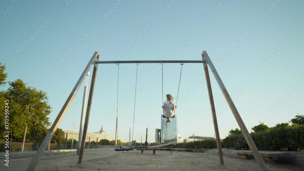 Lonely young woman in white tshirt and jeans swings on swingset on kids playground, flies high in air overlooking city skyline, concept forever youth and happinness Lonely young woman in white tshirt and jeans swings on swingset on kids playground, flies high in air overlooking city skyline, concept forever youth and happinness