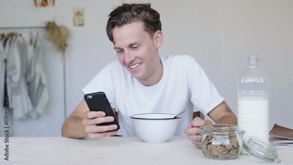 Young attractive man in white pajamas tshirt laughs hard from joke, message or news headline he saw on smartphone on early sunny morning, makes him forget about breakfast Young attractive man in white pajamas tshirt laughs hard from joke, message or news headline he saw on smartphone on early sunny morning, makes him forget about breakfast