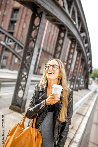 Lifestyle portrait of a stylish businesswoman in leather jacket walking with coffee cup outdoors on the iron bridge