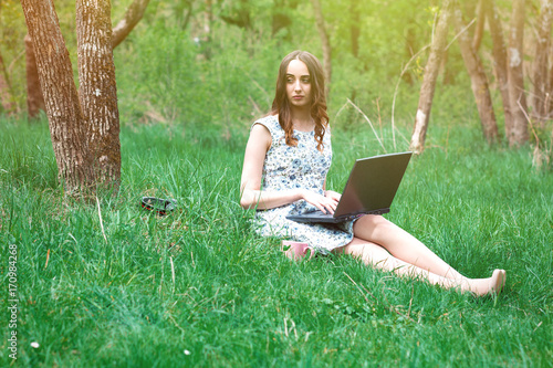 Woman or girl in a dress, with a laptop and headphones, sits on the grass and works with a cup