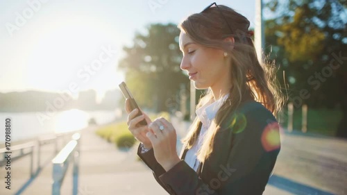 Young Businesswoman Using SmartPhone, Going to Work in the Sunny Morning City. SLOW MOTION. STEADICAM Stabilized Shot. Attractive Professional Business Woman with a cell phone in a city. Lens Flare.  photo