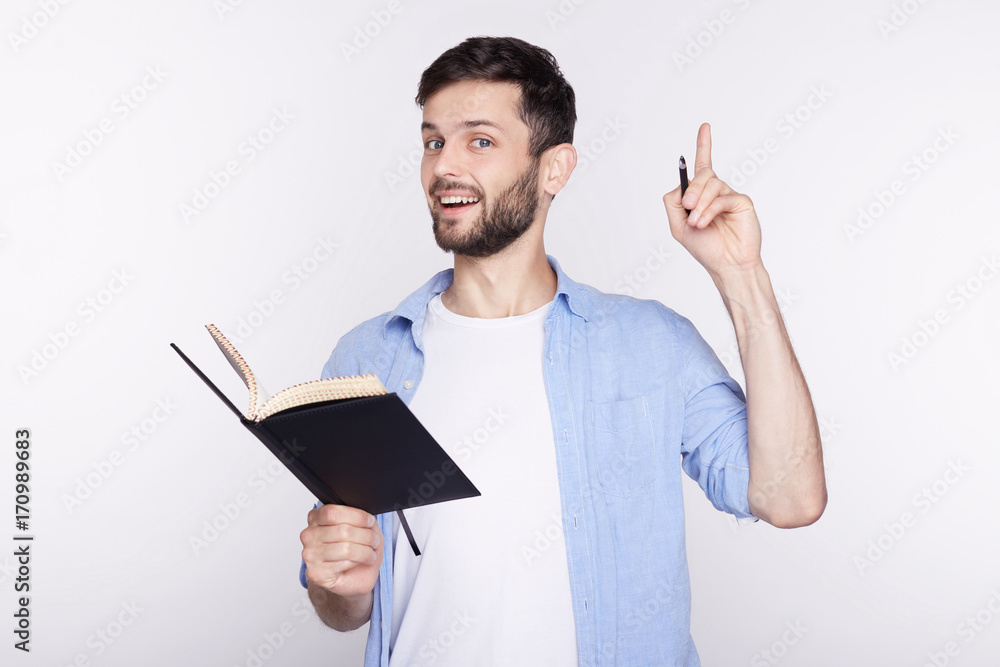 Young amazed Caucasian man with notebook looking at camera with opened mouth showing teeth, pointing his finger at white background with copy space for your advertisement or promotional information. Young amazed Caucasian man with notebook looking at camera with opened mouth showing teeth, pointing his finger at white background with copy space for your advertisement or promotional information.