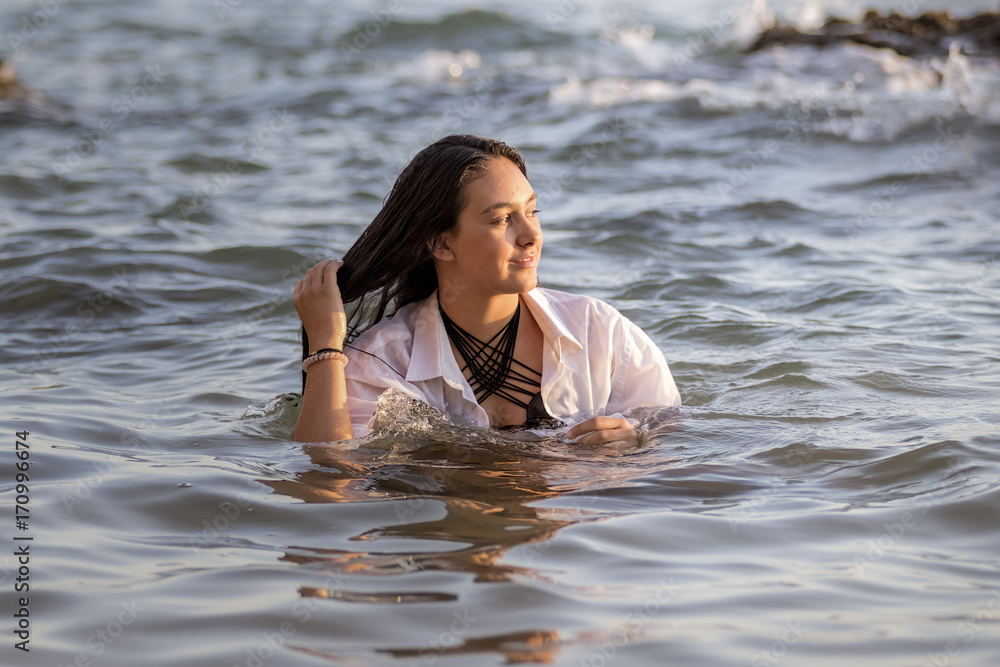 Fototapeta premium Chica joven con camisa blanca bañándose en el mar al atardecer y jugando con su pelo largo moreno.
