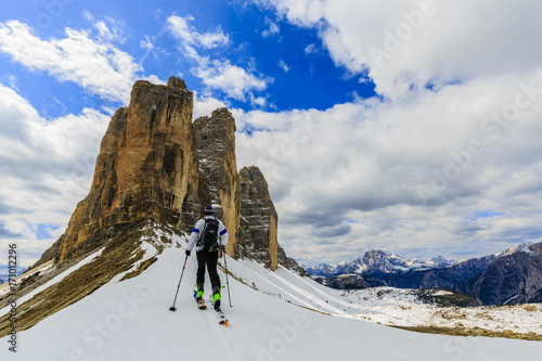 Mountaineer backcountry ski walking up along a snowy ridge with skis in the backpack. In background blue cloudy sky and shiny sun and Tre Cime.