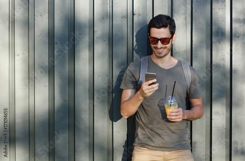 Horizontal outdoor image of young optimistic European man pictured with grey wooden fence behind, looking at screen of phone, browsing and looking through web content while staying in countryside photo