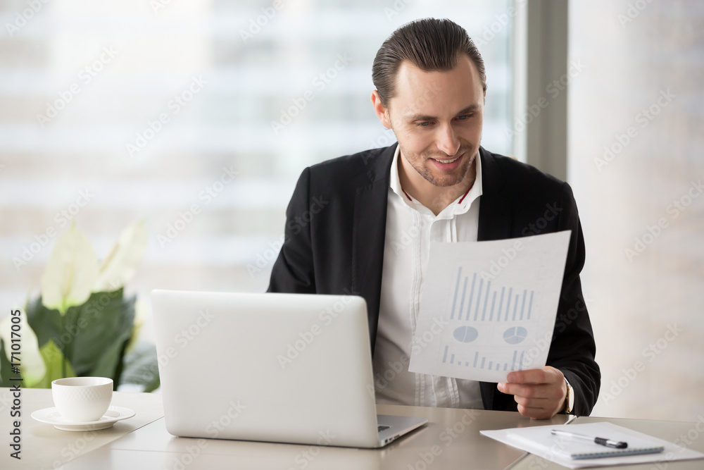 Young cheerful business man sitting in modern office in front of laptop with happy smile, holding financial report document, pleased with successful stock investment profit. Finance success concept. Young cheerful business man sitting in modern office in front of laptop with happy smile, holding financial report document, pleased with successful stock investment profit. Finance success concept.