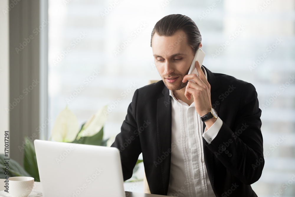 Young handsome businessman sitting at modern office desk looking at laptop screen and making phone call. Remote business negotiations, discussing deals, client consulting, setting up meeting concept. Young handsome businessman sitting at modern office desk looking at laptop screen and making phone call. Remote business negotiations, discussing deals, client consulting, setting up meeting concept.