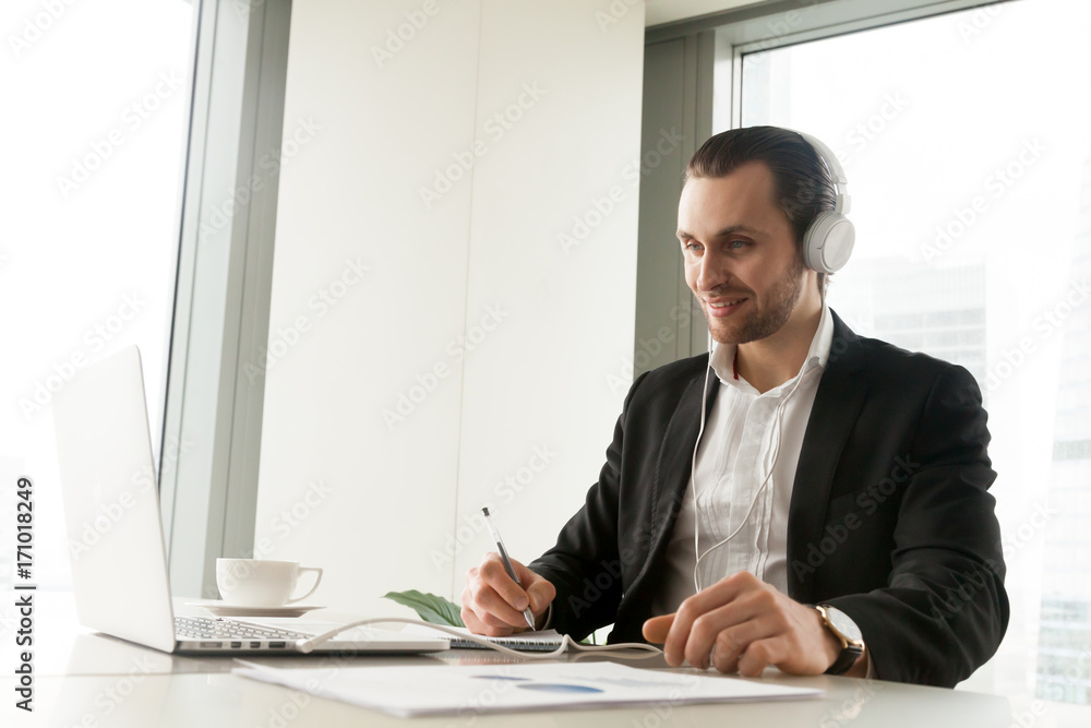 Smiling businessman in headphones taking notes in front of laptop at workplace. Friendly young manager participating in online meeting or conference, remote job interview, learning foreign languages. Smiling businessman in headphones taking notes in front of laptop at workplace. Friendly young manager participating in online meeting or conference, remote job interview, learning foreign languages.