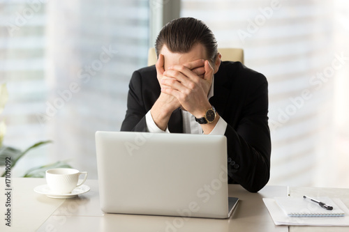 Terrified businessman covering face with hands in front of laptop. Distressed CEO shielding himself from disastrous financial result. Received terrible news, business failure, enormous debt concept.