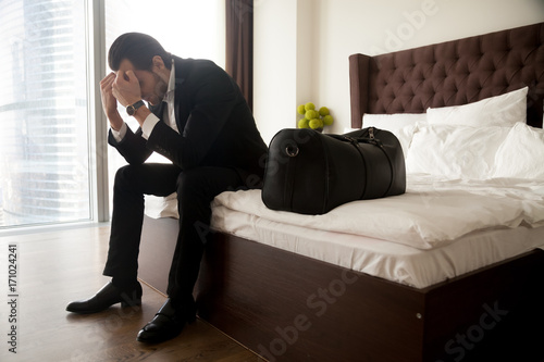 Frustrated young man in formal suit sitting on bed besides luggage bag. Businessman thinking about problems in business or at home, not feeling well, lost job, relationships or work related stress. © fizkes