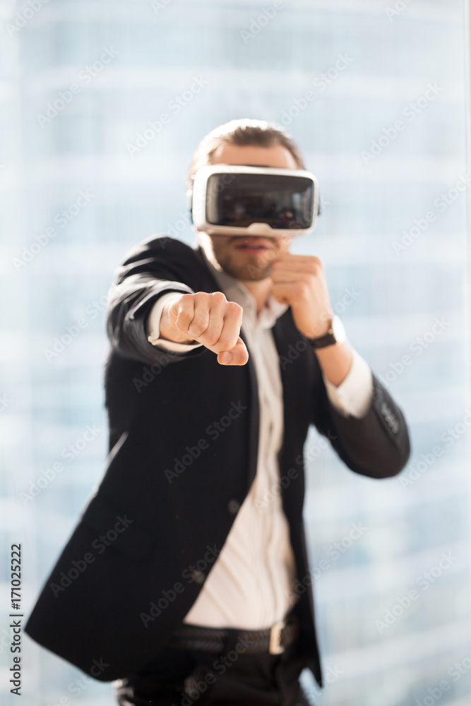 Businessman in VR glasses playing boxing game simulator. Young office worker in formal suit taking break and participating in virtual reality fighting game. Innovation, digital entertainment at work.