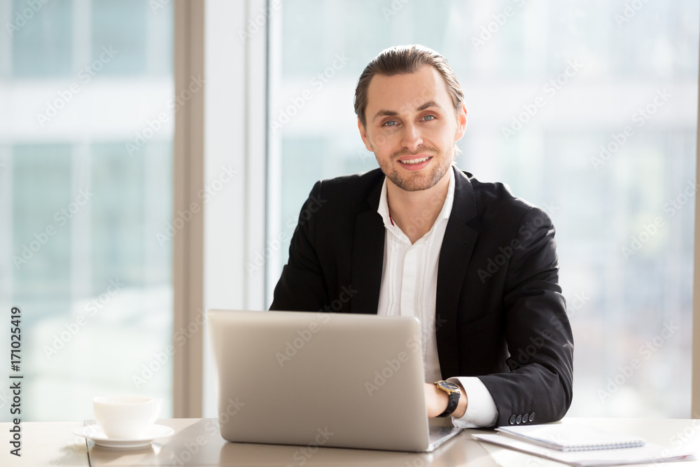 Portrait of handsome smiling businessman sitting in front of laptop at work in modern office. Young entrepreneur enjoying good day at work, friendly company employee working and looking at camera. Portrait of handsome smiling businessman sitting in front of laptop at work in modern office. Young entrepreneur enjoying good day at work, friendly company employee working and looking at camera.