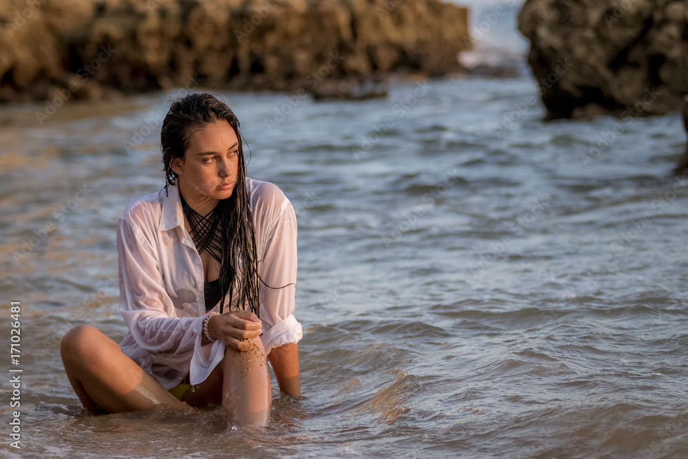 Chica joven con camisa blanca sentada en la playa mojándose con las olas  del mar al atardecer. Stock Photo | Adobe Stock
