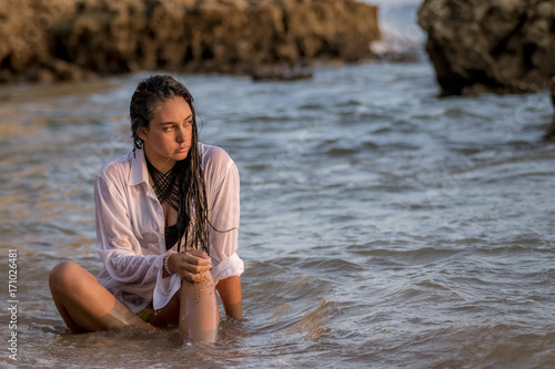 Fototapeta Naklejka Na Ścianę i Meble -  Chica joven con camisa blanca sentada en la playa mojándose con las olas del mar al atardecer.