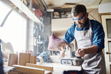Confident bearded carpenter wearing apron and safety goggles working with belt sander, interior of messy workshop on background
