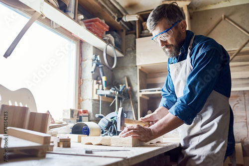 Fotografia, Obraz Bearded middle-aged craftsman wearing safety goggles and apron hammering nails i