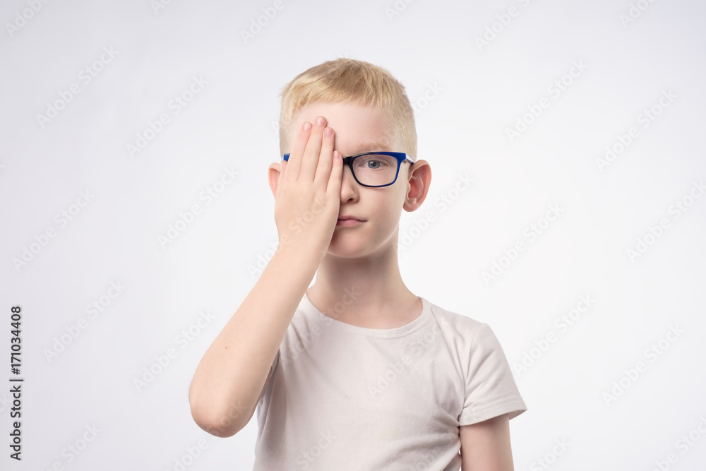 Caucasian blond child reviewing eyesight closing half of face with hand.