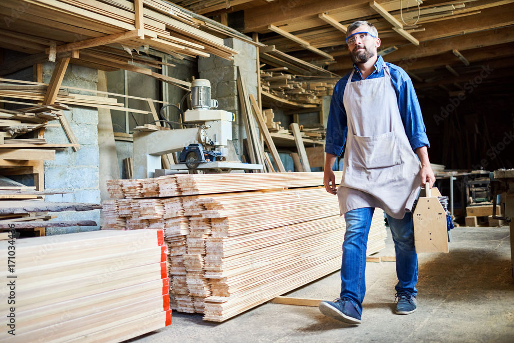 Full length portrait of confident bearded carpenter wearing apron and safety goggles walking along workshop with tools kit in hand Full length portrait of confident bearded carpenter wearing apron and safety goggles walking along workshop with tools kit in hand