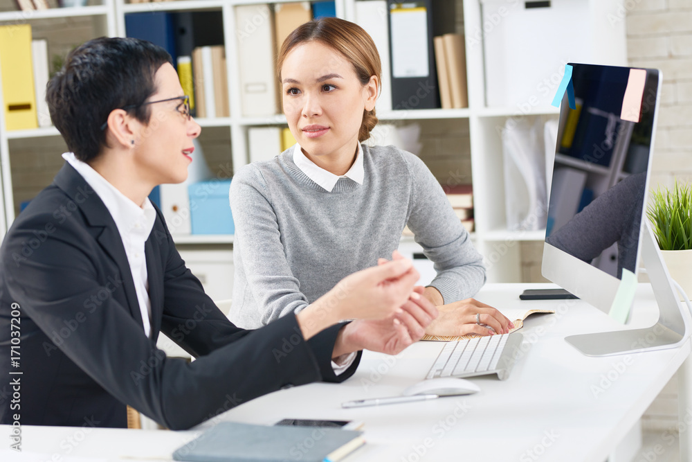 Pretty middle-aged white collar worker presenting her point of view concerning promising project to young Asian colleague while sitting at desk in modern open plan office Pretty middle-aged white collar worker presenting her point of view concerning promising project to young Asian colleague while sitting at desk in modern open plan office