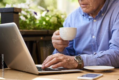 Closeup image of unrecognizable senior man using laptop computer in cafe outdoors holding coffee cup
