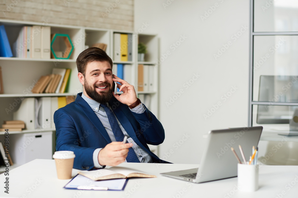 Portrait of successful bearded businessman speaking by phone looking at camera while working at desk in light office Portrait of successful bearded businessman speaking by phone looking at camera while working at desk in light office