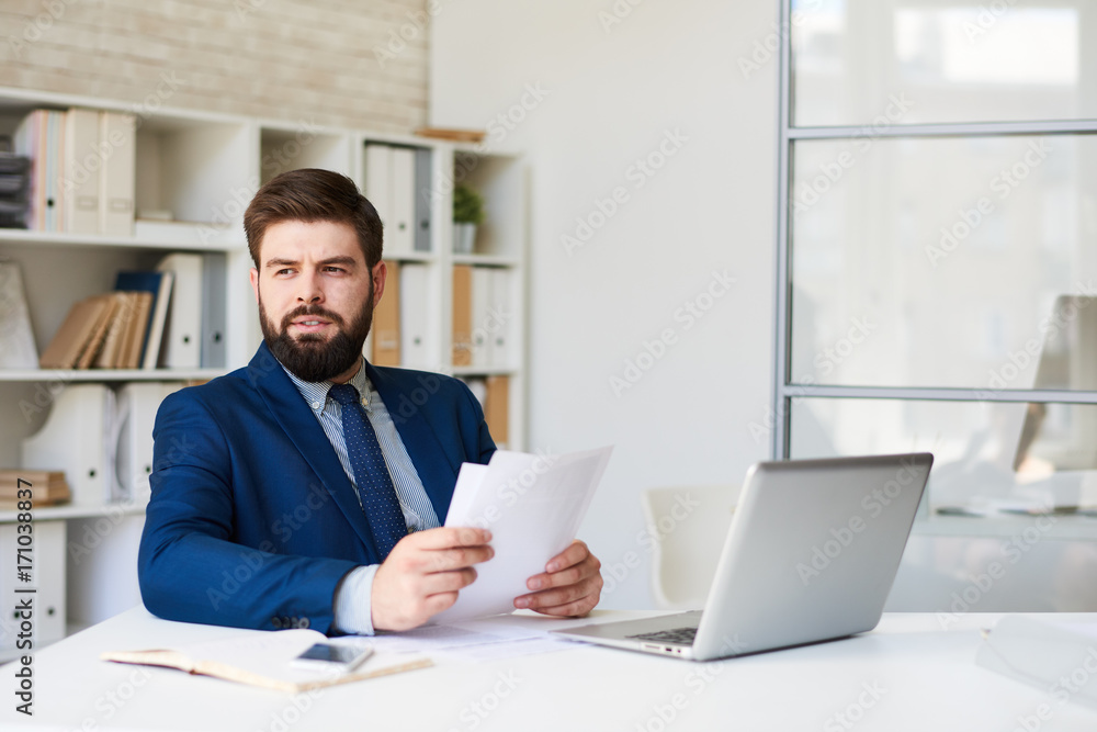Portrait of successful  bearded businessman  looking away pensively while working at desk in modern office
