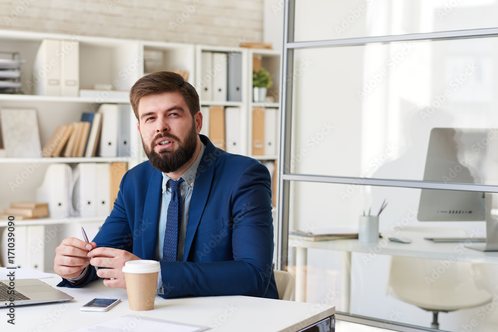 Portrait of successful bearded businessman looking away thoughtfully while working at desk in modern office Portrait of successful bearded businessman looking away thoughtfully while working at desk in modern office