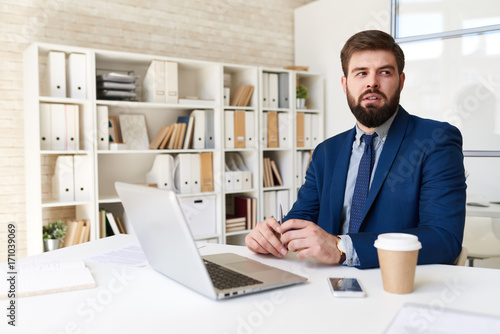 Portrait of successful  bearded businessman looking away thinking while working with laptop  in modern office