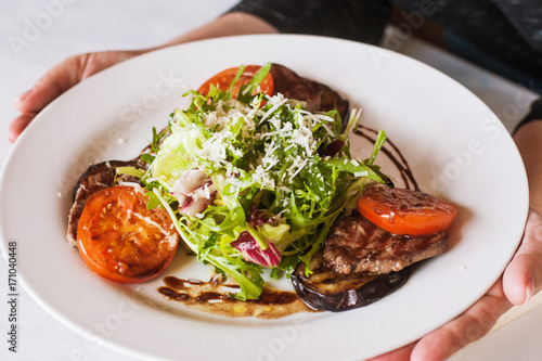 Medallions with grilled vegetables on plate bringing to customer. Meat, tomatoes, eggplant and green salad with grated cheese, serving in restaurant