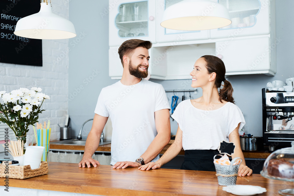 Couple of young small business owners looking at each other while standing behind counter in their cafe Couple of young small business owners looking at each other while standing behind counter in their cafe