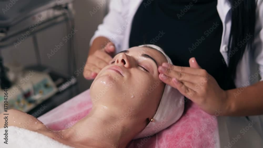 Closeup view of a professional cosmetologist's hands making face and neck massage in spa salon. Young woman is lying on the couch during cosmetic face procedure. Beauty, healthy and youth concept Closeup view of a professional cosmetologist's hands making face and neck massage in spa salon. Young woman is lying on the couch during cosmetic face procedure. Beauty, healthy and youth concept