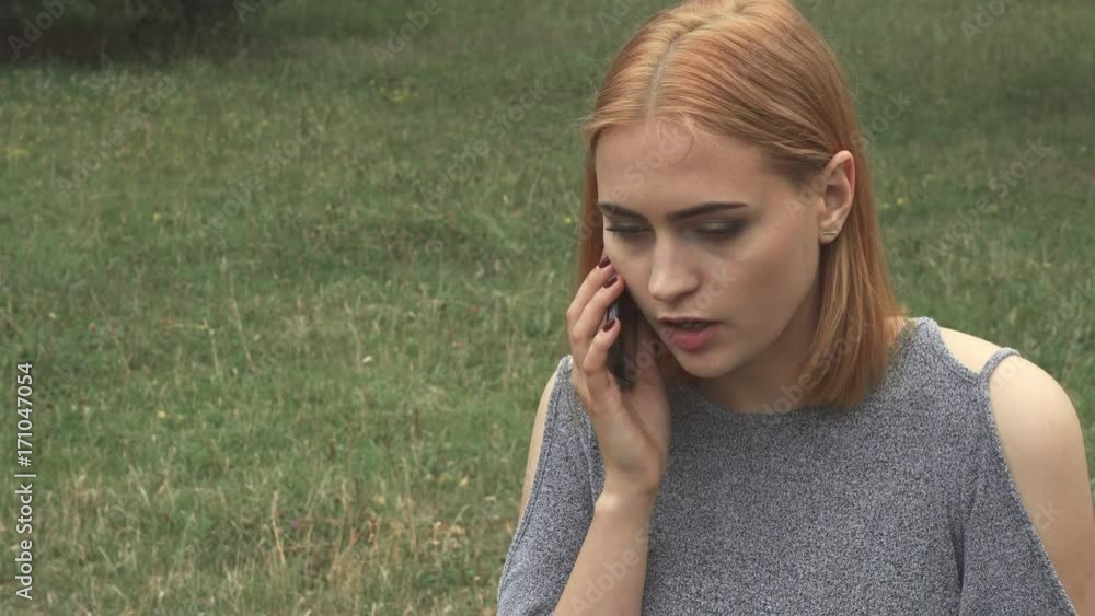 Blonde talking on phone being upset. Then she finishes talking ang massage her temples. Woman sitting on grass in park Blonde talking on phone being upset. Then she finishes talking ang massage her temples. Woman sitting on grass in park