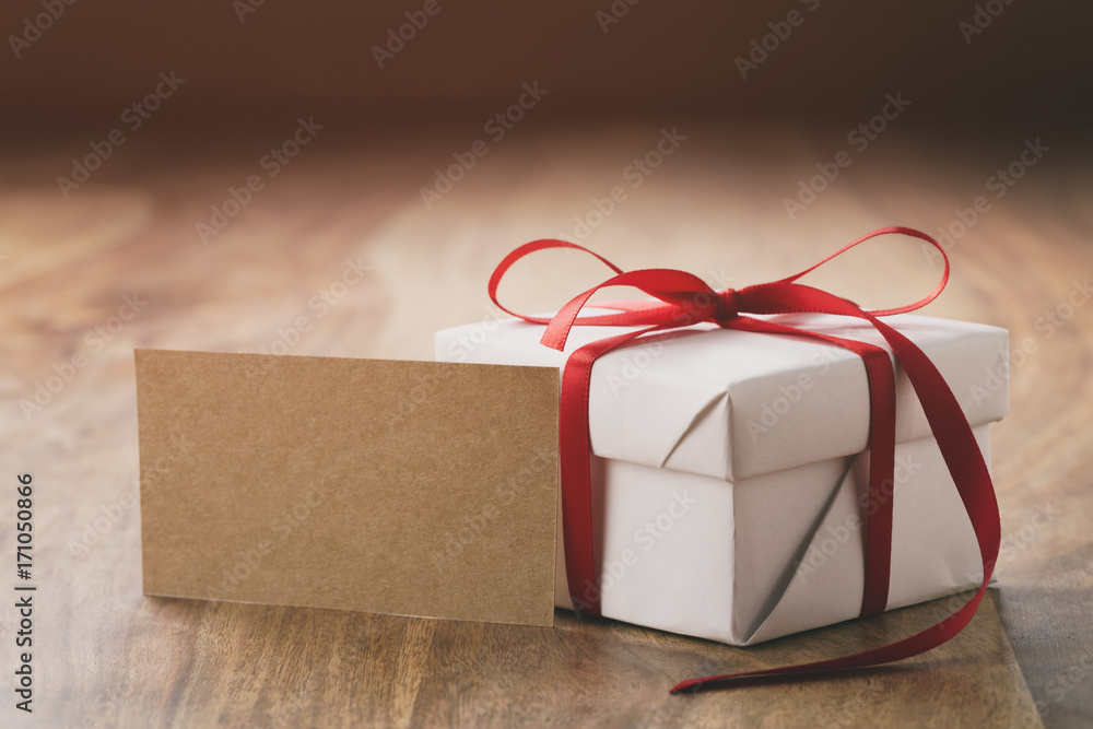 white gift box with thin red ribbon bow on old wood table with paper card  Stock Photo