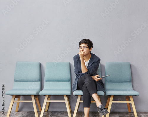 Picture of attractive fashionable young female designer wearing trendy clothes and eyeglasses sitting on chair in hall, keeping legs crossed, feeling nervous before job interview, biting her nails photo