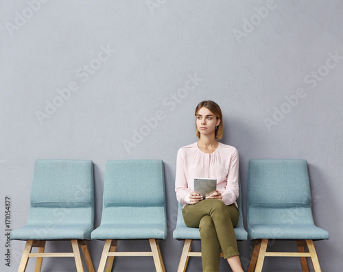 Portrait of confident serious young European female sitting on chair in waiting room with electronic tablet, setting her mind up before job interview or meeting with potential business partners photo