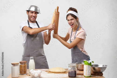 Cookers competitors participate in contest, fight with rolling pins, have angry expressions. Professional male and female rivals on kitchen, competiting who cooks better. Culinary competition photo