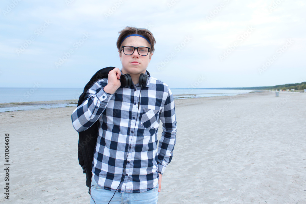 A boy is a teenager (a schoolboy or a student) holding a jacket, wearing glasses, wearing a shirt, headphones on the sea.