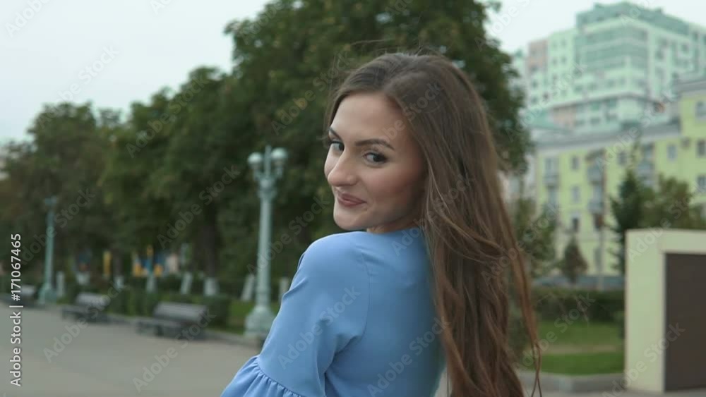 Young beautiful woman in a blue dress walking the river embankment. She is smiling, posing for the camera, her hair blowing in the wind, looking very happy. Young beautiful woman in a blue dress walking the river embankment. She is smiling, posing for the camera, her hair blowing in the wind, looking very happy.