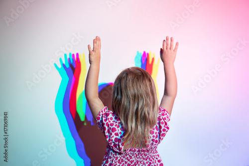 Little girl stands against the wall with her arms raised with her back to the camera. On the wall is a diffraction shadow photo