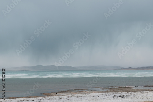 panoramic view of watery surface of lake with snow on coast and storm clouds in sky 