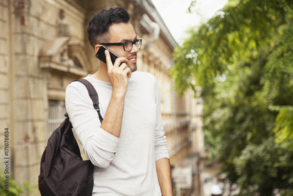 Young excited happy male tourist with a backpack talking with friend on phone in old European town (communication, tourism, vacation, holiday concept)