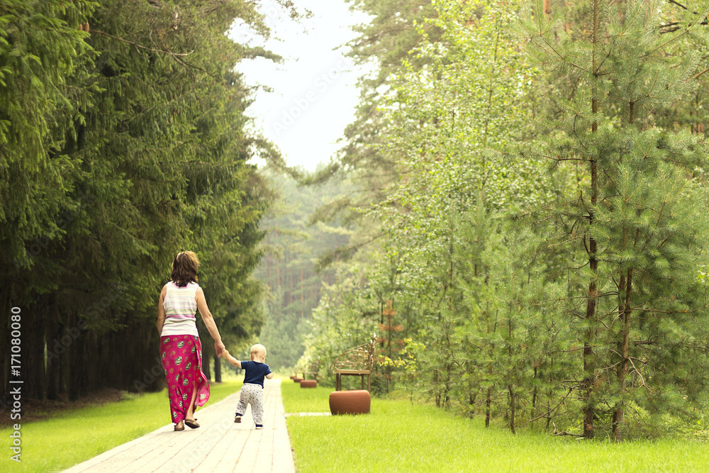 Fototapeta premium Mom and son on the walk in the park. Back view of walking family. First independent steps of one year toddler.