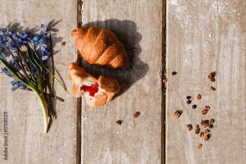Delicious breakfast on wooden background. Sweet croissant and granola with spring blue snowdrops nearby. Morning fresh and tasty meal. Food photography top view