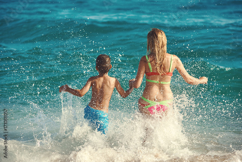 Caucasian mom and son running into the sea water. Back view.